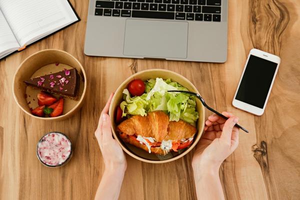 desk with laptop, phone, bowl with slice of cake and bowl with sandwich and salad and person's arms holding bowl and fork
