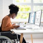 photo of Black woman wearing an orange shirt and black pants in wheelchair working on computer